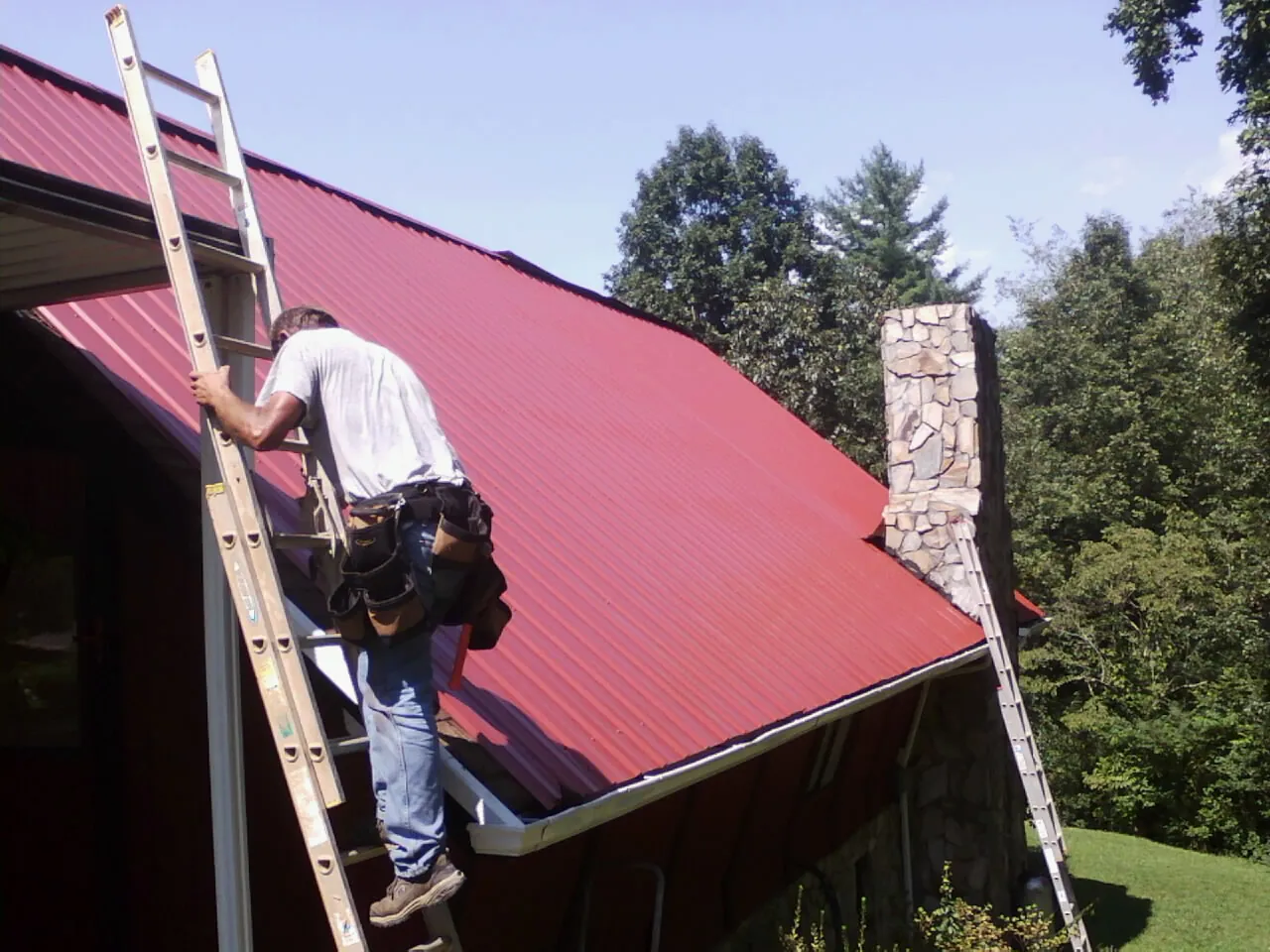 Roofer on a ladder beside a red metal roof and stone chimney — Soffit & Fascia Repair by Roofing Brothers in Phippsburg