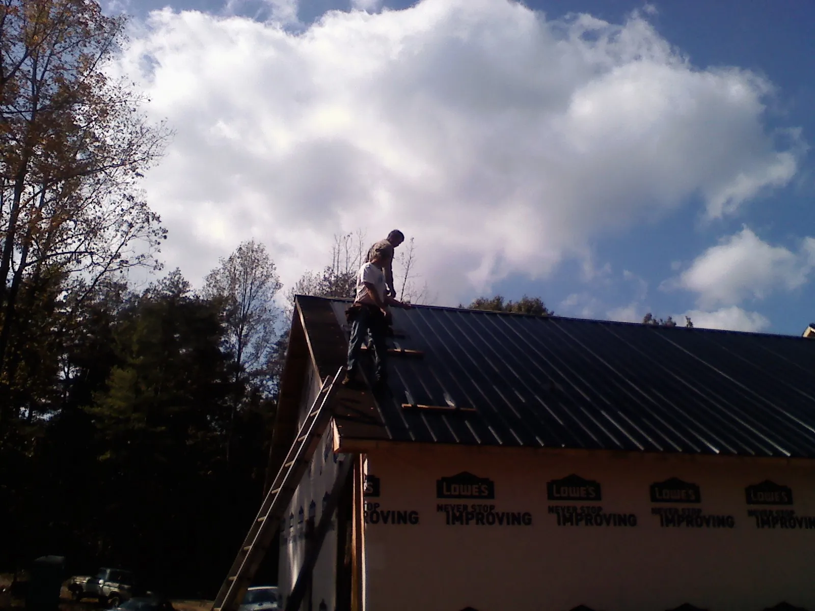 Roofer fastening dark metal panels high against a cloudy sky, from a Roof Inspection job in Phippsburg